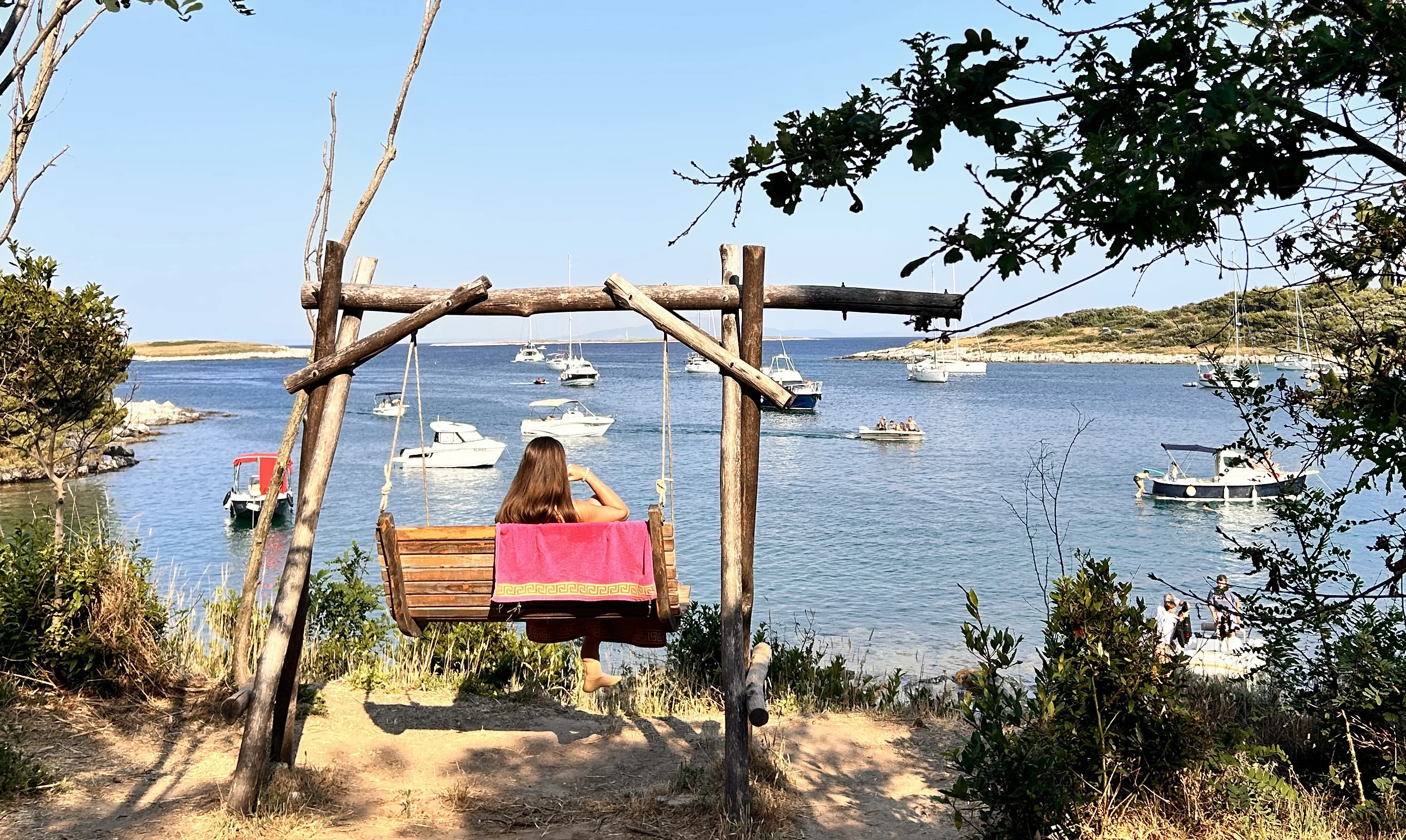 osoba siedząca tyłem na dużej, drewnianej huśtawce ustawionej na klifie nad zatoką. Huśtawka jest zrobiona z grubych gałęzi i desek. Na oparciu huśtawki wisi kolorowy ręcznik. Przed osobą rozciąga się spokojna, błękitna woda z kilkoma łodziami i jachtami. Wokół zatoki rośnie niska roślinność i krzewy. Słońce świeci jasno, tworząc letnią, spokojną atmosferę