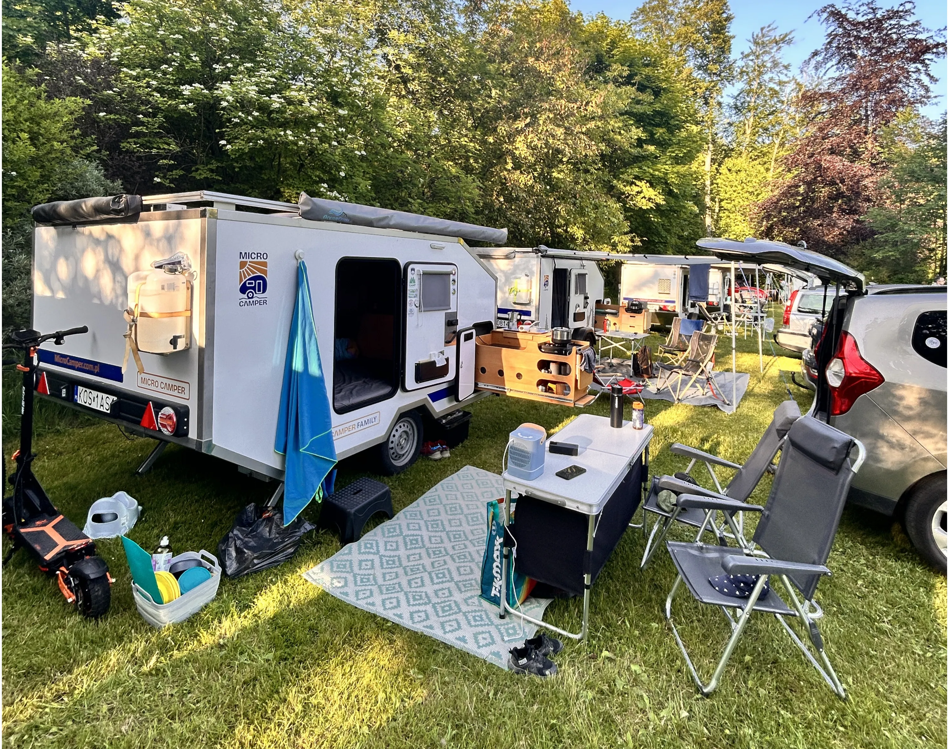 Kemping. pole kempingowe z przyczepami. Na pierwszym planie stoi mała biała przyczepa kempingowa z otwartymi schowkami i rozwieszonym ręcznikiem. Obok znajduje się składany stolik z kubkami i drobnymi rzeczami oraz dwa turystyczne krzesła. Wokół rozstawiono różne rzeczy: hulajnoga, pojemniki z rzeczami i mata piknikowa. W tle widać kilka podobnych przyczep i samochodów.